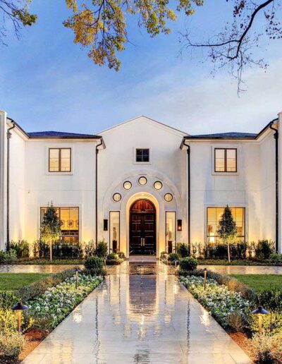 Modern white stucco house with symmetrical design featuring a central arched doorway, flanked by large trees, and a pathway leading to the front door at dusk.