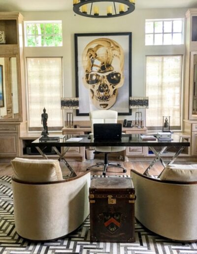 A stylish home office featuring two cream chairs, a desk with a computer, bookshelves, ornate cabinetry, and a large skull artwork above the desk.