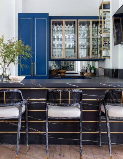 A modern kitchen with blue cabinets, black marble countertops, and rattan bar stools set against wood flooring.