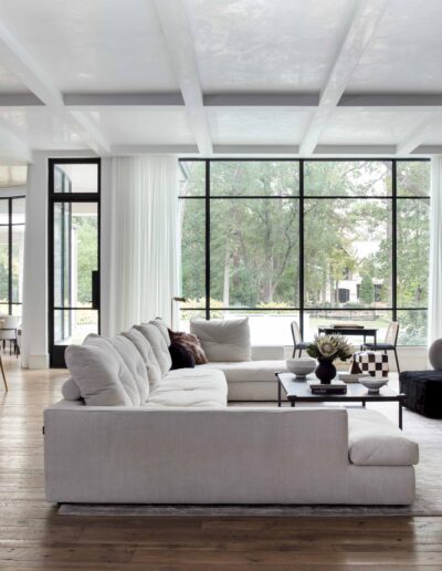 A spacious, modern living room with large windows, a white sofa, dark armchairs, and a fireplace.