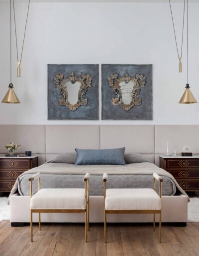 Elegant bedroom featuring a neat bed with blue pillows, two wall art pieces above, antique-style bedside tables, and brass hanging lamps.