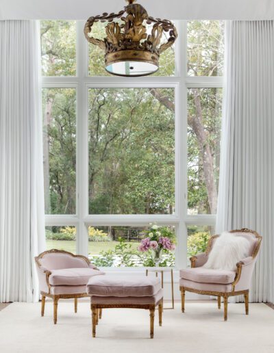 Elegant living room interior with two vintage chairs by a large window overlooking a garden, adorned by a floral chandelier and white drapes.
