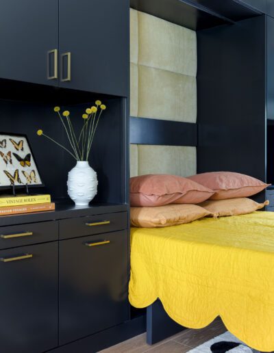 A modern bedroom with a yellow bedspread, black cabinetry, and a unique white vase with green stems on a shelf.