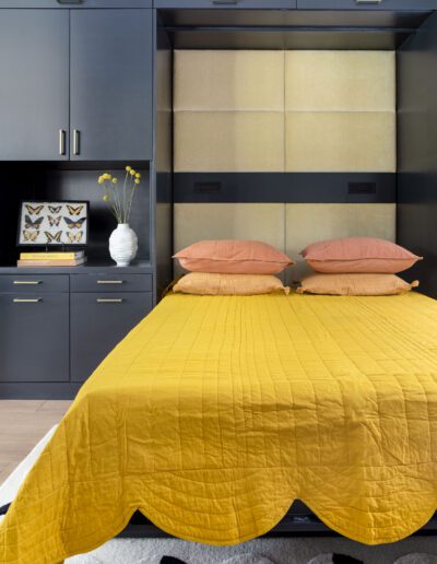 Modern bedroom with a mustard yellow bedspread, peach pillows, grey cabinets, and an integrated wall padding design.