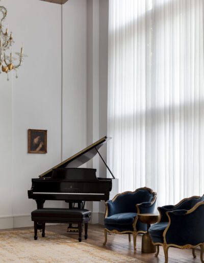 A grand piano in a luxurious room with sheer curtains, two ornate blue velvet chairs, a gold chandelier, and a small painting on the wall.