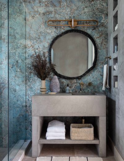 A stylish bathroom with a concrete sink, a round mirror, and textured blue walls, decorated with a dried plant and accessories.