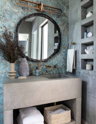 Elegant bathroom featuring a concrete sink, round mirror, and decorative items on storage shelves against a textured blue wall.